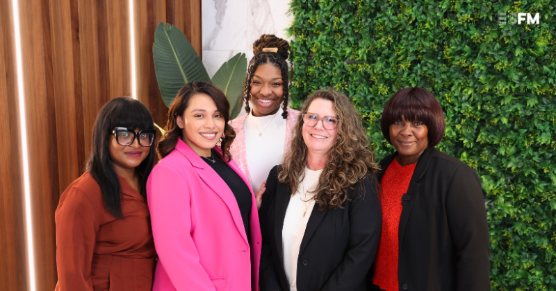 Panelists from the women's history month discussion pose in front of a bright backdrop of polished wood and greenery.